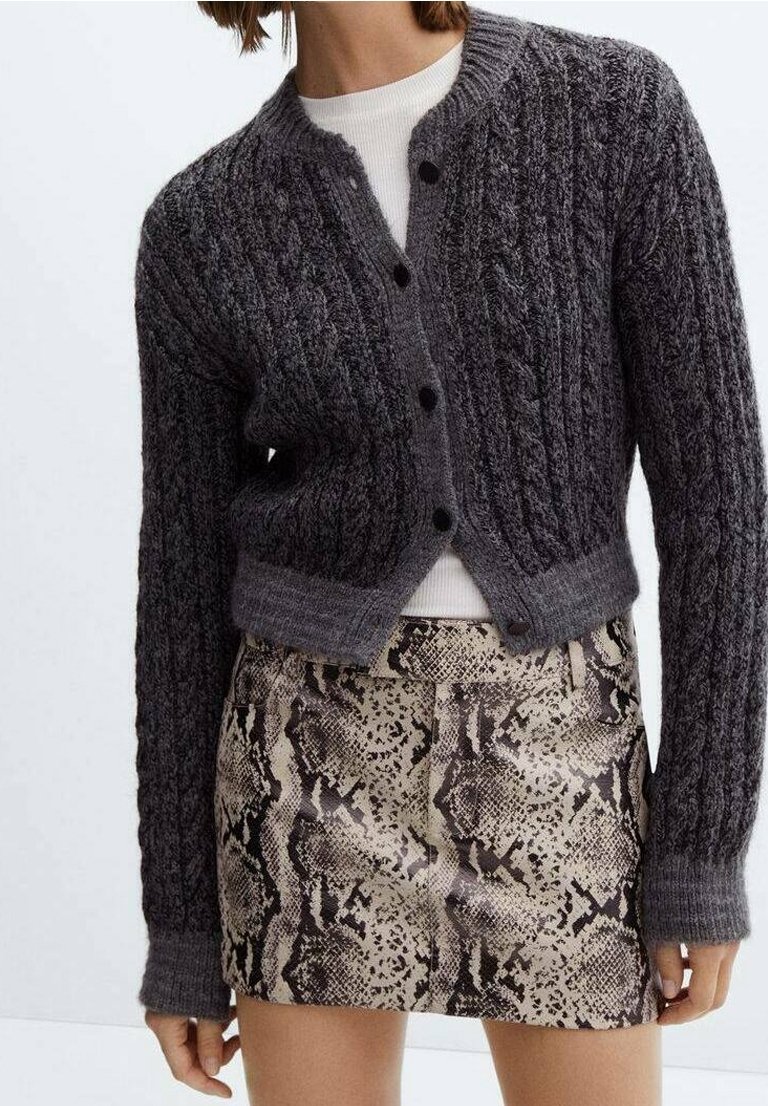 Mørkegrå kabelstrik cardigan med knaplukning, cropped design. Båret over en cremefarvet top og parret med en mønstret slangeskindsprint nederdel.