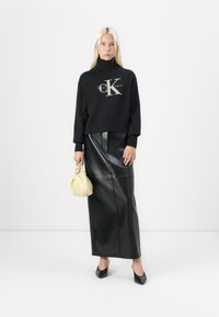 Zwarte cropped turtle neck trui met 'Calvin Klein' logo, gecombineerd met een lange zwarte leren rok en een gele stoffen handtas. Zwarte schoenen.
