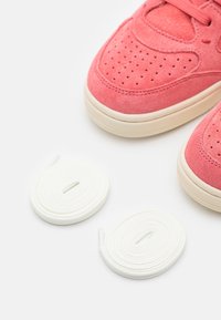 Rosa mockasneakers med perforerade detaljer och vita gummisulor, tillsammans med två vita skosnören som är ringlade på en vit yta.