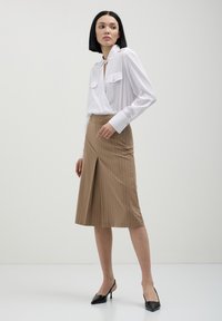 Blusa bianca a maniche lunghe con due tasche frontali e scollo a V, abbinata a una gonna marrone a righe verticali con spacco frontale e vita aderente.