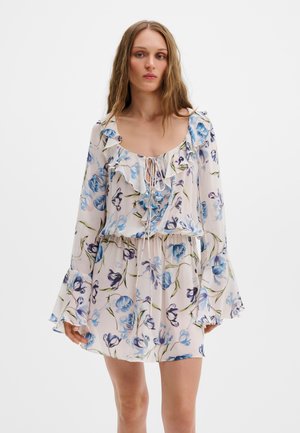 Žena v bielych kvetinových šatách s modrými a fialovými kvetmi, s dlhými zvonovými rukávmi, volánikovým výstrihom a zaväzovaním na hrudi.