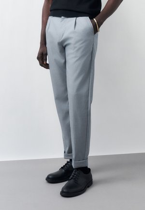 Mężczyzna ubrany w jasnoszare, szyte na miarę spodnie z mankietami, czarne sznurowane buty, czarną koszulę i złotą bransoletkę, stojący na jednolitym tle.