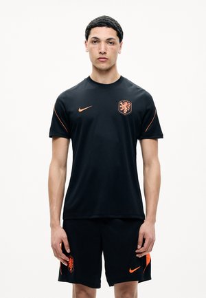 Junger Mann steht frontal, trägt schwarze Sportbekleidung mit orangefarbenen Nike-Logos und Löwenwappen auf Shirt und Shorts.