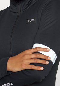 Svart zip-up jacka i stretchigt material, med en vit detalj på ärmen och en GORE-logotyp på bröstet.