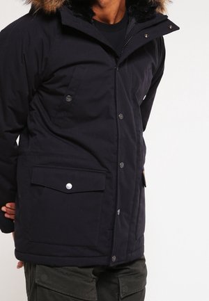 Hombre con un parka de invierno negro con capucha con borde de piel, botones de presión delanteros, bolsillos con solapa y pantalones cargo oscuros.