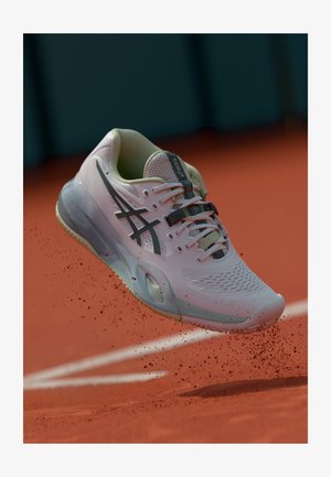 Jasnoróżowy but sportowy z siateczkową cholewką, czarnymi akcentami i wzorzystą podeszwą, pokazany w powietrzu nad czerwoną glinianą powierzchnią, z cząstkami kurzu.