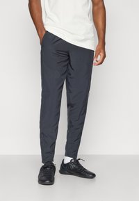 Slim fit, sötétszürke melegítők oldalsó csíkokkal, sima textúrával, rugalmas derékrésszel és cipzáras zsebekkel. Fekete sportcipővel viselve.