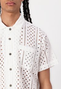 White short-sleeve shirt made of embroidered cotton fabric, featuring cut-out floral patterns, a chest pocket, and button closure.