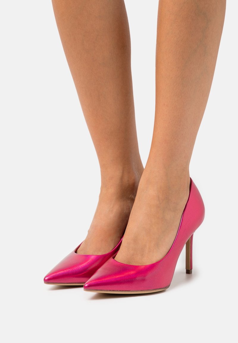 Pieds d'une femme portant des chaussures à talons hauts brillantes, à bout pointu, rose vif sur un fond clair uni.