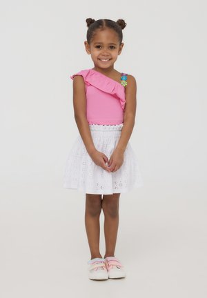 Top rosa arricciato a spalla unica con dettaglio floreale, abbinato a una gonna bianca a strati in pizzo. Sneakers pastello morbide completano l'outfit.