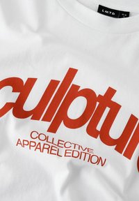 Vit bomulls t-shirt med stor röd text som säger "skulptur KOLLEKTIV KLÄDUTGÅVA." Slät textur, rundad halsdesign.