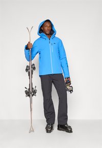 Jaqueta impermeável azul brilhante com capuz, calças de esqui pretas e botas de esqui pretas. Segurando esquis e óculos de esqui.