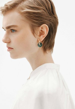 Pendientes circulares verdes con un acento dorado, con un diseño en capas. Modelo con cabello corto lleva una blusa blanca con cuello frente a un fondo minimalista.
