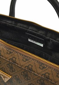 Interior de bolso marrón con estampado, forro negro, cremallera dorada y una etiqueta negra que muestra "GUESS ECO".