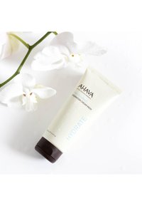 Mascarilla hidratante AHAVA en un tubo beige con tapa marrón, con texto azul. Rodeada de flores de orquídea blancas y gotas sobre un fondo blanco.