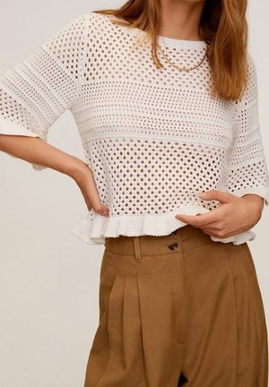 Woman wearing a white crocheted short-sleeve top with ruffled hem and brown high-waisted pleated pants, posing with one hand on waist.