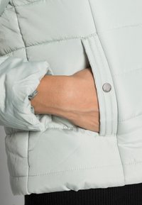 Jasnoszara zielona kurtka puchowa o fakturowanym wykończeniu, z boczną kieszenią i ozdobnym guzikiem, z marszczonym mankietem i przeszywanymi szwami.