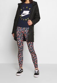 Svart vattentät jacka, blå grafisk sweatshirt, blommönstrade leggings och svarta sneakers med vita sulor.