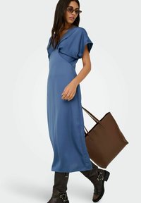 Niebieska sukienka do kolan z gładkiego materiału, z plisowanym dekoltem w kształcie litery V i krótkimi rękawami. Noszona z brązową torebką i wysokimi czarnymi botkami.