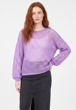 Rødhåret kvinde iført en løs, lavendel åbenstrikket sweater over en matchende top og et sort knælange nederdel med frontslids.
