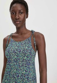 Blumenmusterkleid mit lila Grundfarbe, das grüne und blaue Muster aufweist. Dünne Träger sind an den Schultern gebunden, und der Stoff hat eine glatte Textur.