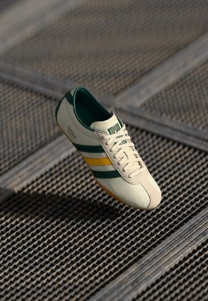 White sneaker with green and yellow stripes, Adidas Tokyo branding, light brown sole, resting on textured metal grate.
