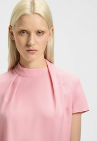 Top rosa a maniche corte con scollatura alta e morbide pieghe al colletto. Tessuto dal tocco liscio, design minimalista e lucentezza sottile.