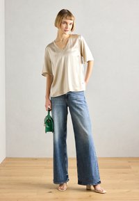 Beige, kortermet satengbluse med V-hals, kombinert med brede blå denimjeans. Holder en liten grønn håndveske. Lys tregulv.