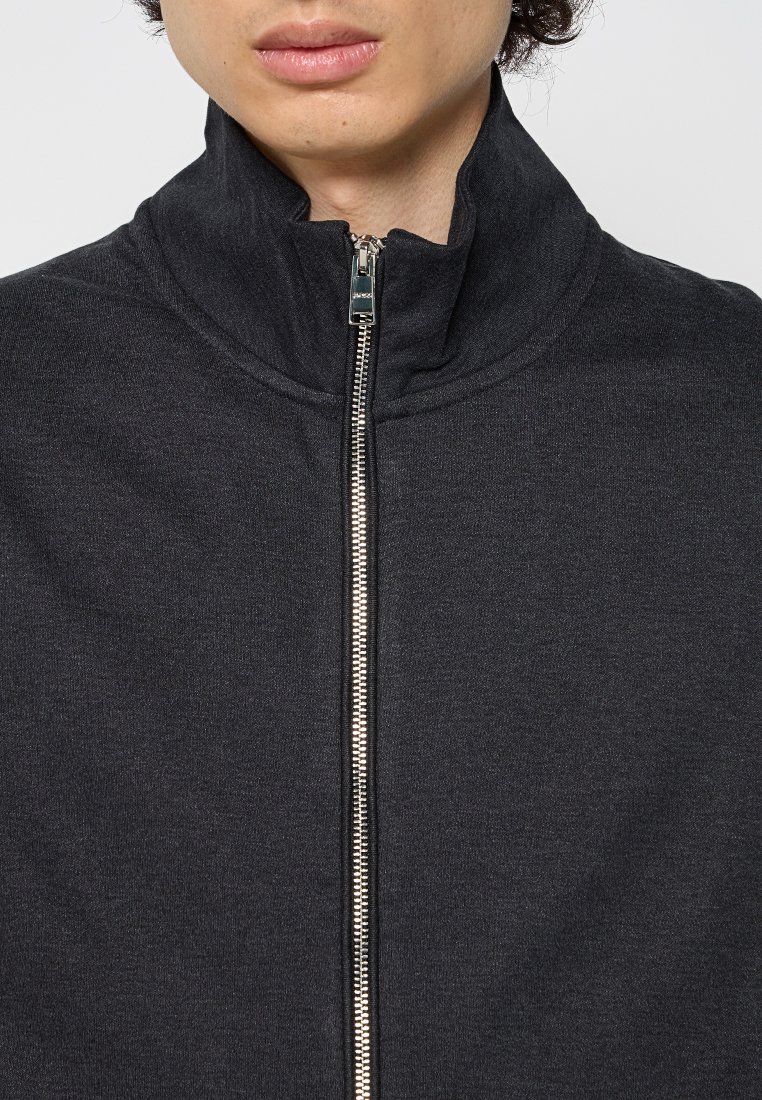 Persoon die een donkergrijze ritsjack draagt met de rits volledig gesloten tot een hoge kraag, waarbij alleen de lippen en kin zichtbaar zijn.