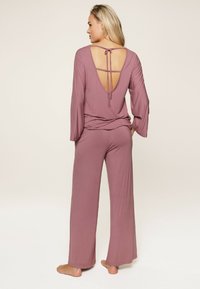 Set di abbigliamento loungewear in tinta mauve, comodo e morbido, con maniche lunghe, apertura profonda sulla schiena e dettaglio di lacci. Tessuto morbido, pantaloni a gamba larga, accenti minimal.