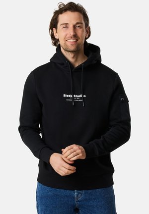 Zwarte hoodie met het logo "Slade Studios" in het wit, voorzien van een voorzak in kangoeroestijl en geribbelde manchetten. Denim jeans maken de outfit compleet.