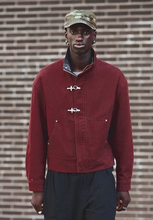 Homme portant une veste rouge avec des fermetures à brandebourgs, un pantalon noir, une casquette camouflage et des boucles d'oreilles en argent en forme de créoles, debout devant un mur en briques.