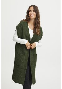 Hosszú, nyitott elülső mellény sötétzöld bordás textúrában, rövid ujjal és két elülső zsebbel. Fehér felső és fekete nadrág fölött viselve.