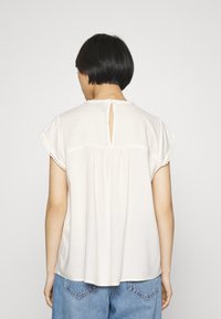 Donna con capelli neri corti che indossa una blusa bianca corta e larga con un'apertura a chiave sulla schiena e jeans di denim azzurri.