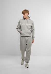 Felpa grigia con cappuccio e pantaloni della tuta abbinati, realizzati in un materiale morbido, caratterizzati da una tasca frontale e testo del logo in bianco. Le sneakers completano l'outfit.