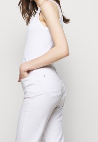 Bílý tílko spárované s bílými džíny. Džíny mají hladkou texturu, zadní kapsy a jemné švy.