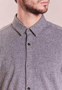 Nærbillede af en mand iført en struktureret grå skjorte med knapper, som viser krave og øvre bryst, med trimmet skæg og neutral baggrund.