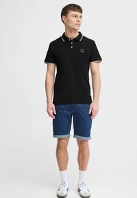 Svart polo skjorte med kontrasterende hvit krage og mansjettkanter, med en liten sirkulær logo, sammen med blå denimshorts og hvite sokker.