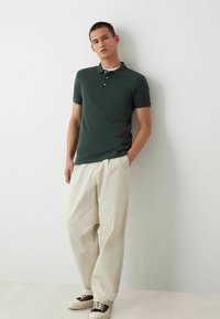 Camisa polo verde oscuro con cuello, tapeta de tres botones y logotipo en la manga izquierda, combinada con pantalones sueltos de color crema y zapatillas.