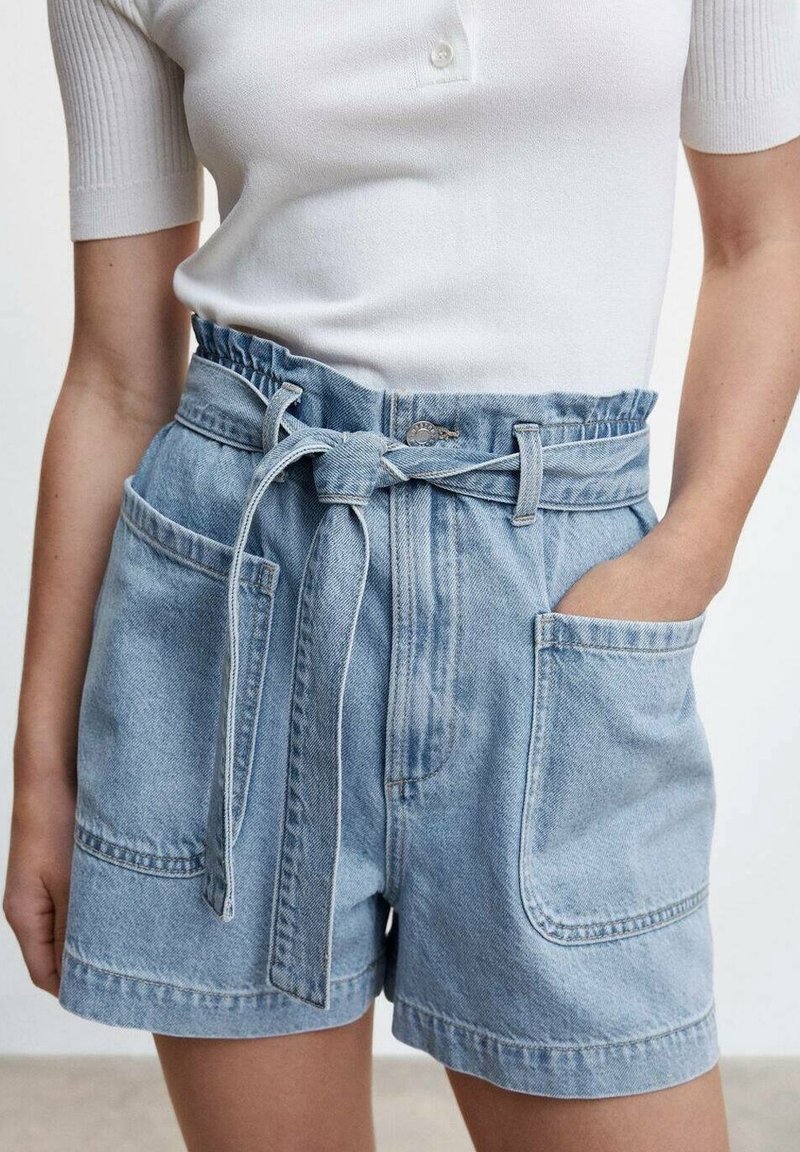 Femme portant un short en jean taille haute bleu clair avec une ceinture nouée et de grandes poches avant, associé à un top blanc côtelé à manches courtes.
