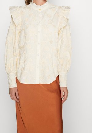Blouse blanche avec des motifs floraux et de cœurs brodés, détails à volants aux épaules et fermeture par boutons. Assortie à une jupe en satin marron.
