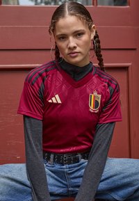 Burgundijas futbola krekls ar melniem akcentiem, ar dimanta rakstu, zelta logotipu un v-veida kakla dizainu. Apvienots ar pelēkiem garajiem piedurknēm un džinsiem.