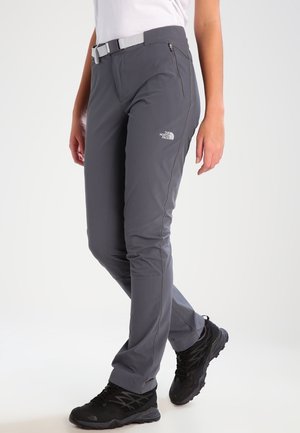 Kobieta nosząca szare spodnie outdoorowe The North Face z zasuwaną kieszenią i białym paskiem, w połączeniu z czarnymi butami trekkingowymi, stojąca na białym tle.