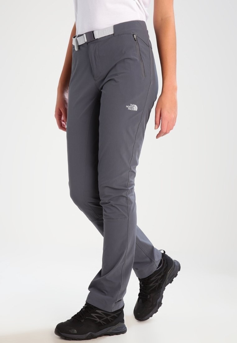 Femme portant un pantalon d'extérieur gris The North Face avec poche zippée et ceinture blanche, associée à des chaussures de randonnée noires, se tenant sur un fond blanc.