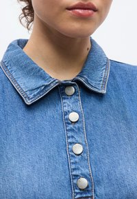 Farmere ing strukturált gallérral, kék árnyalatban, öt fém kapocsgombbal az elején. Simább textúra látható varrási részletekkel.