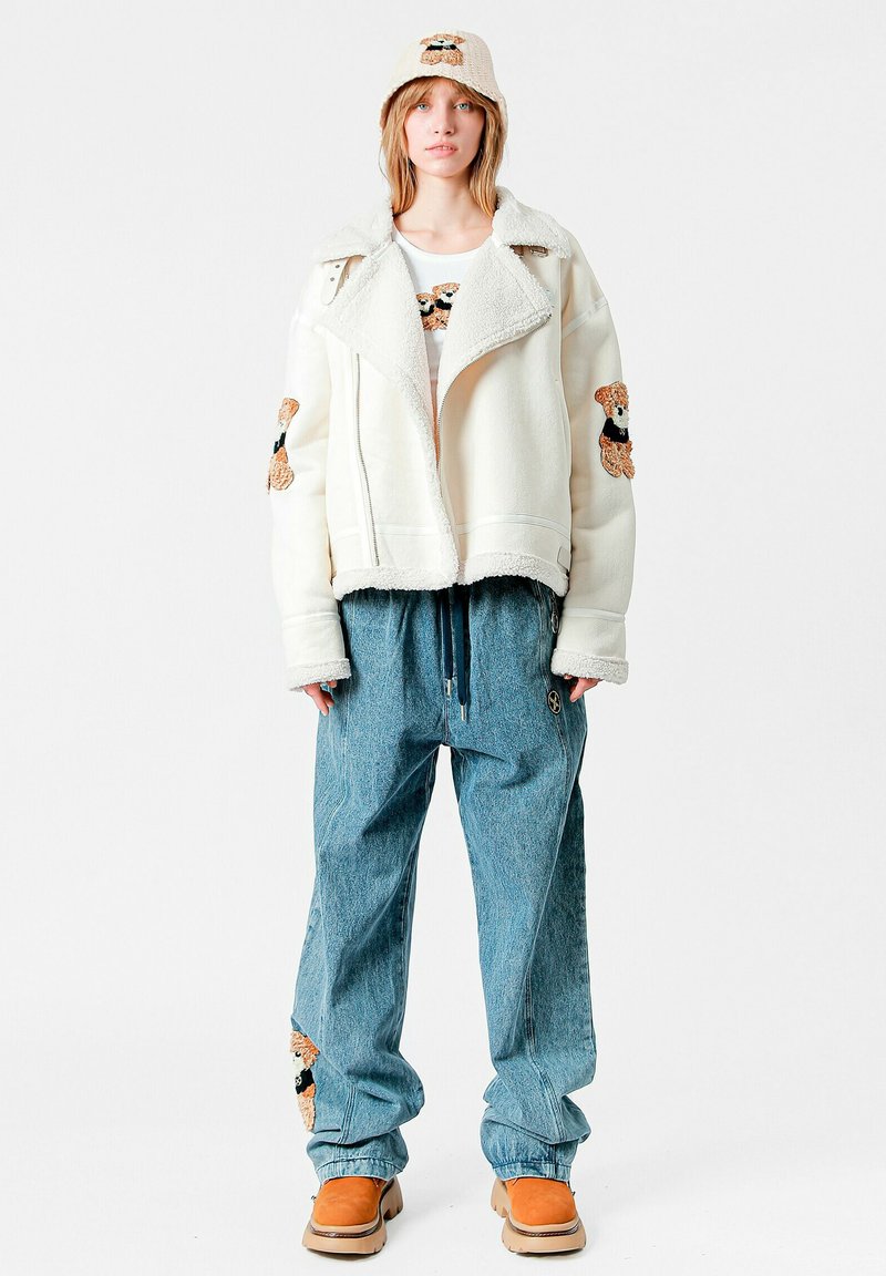Chaqueta de forro polar blanca con parches de osito de peluche, pantalones vaqueros azules oversize con bolsillos y un sombrero beige a juego. Botas marrones completan el conjunto.