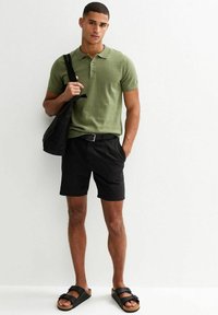 Olive green polo z krótkim rękawem, czarne szorty i czarne sandały. Model trzyma czarną torbę na ramię. Wykonany z bawełny, o prostym kroju, luźny fason.