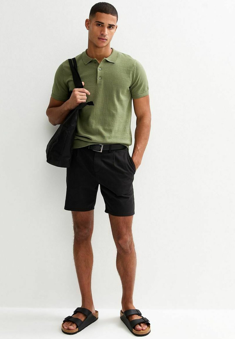Olive green polo z krótkim rękawem, czarne szorty i czarne sandały. Model trzyma czarną torbę na ramię. Wykonany z bawełny, o prostym kroju, luźny fason.