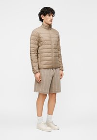 Bézs színű steppelt téli dzseki magas gallérral, steppelt textúrával és elülső cipzárral, bézs mintás rövidnadrággal és fehér tornacipővel párosítva.