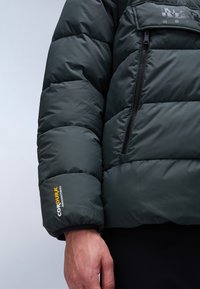 Zbliżenie na rękaw ciemnozielonej kurtki pikowanej z logo "CORDURA Advanced Fabrics" oraz dłonią widoczną na jednolitym tle.